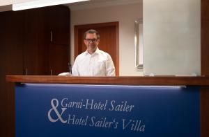 Garni-Hotel Sailer & Hotel Sailer´s Villa في روتويل: رجل يقف خلف مكتب في غرفة