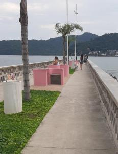 um calçadão com bancos cor-de-rosa e pessoas caminhando ao longo da água em Apartamento pé na areia Praia do Gonzaguinha. em São Vicente