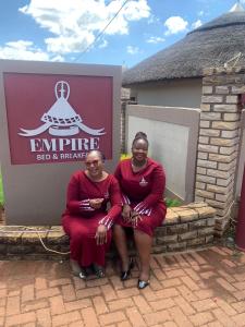 dos mujeres vestidas de rojo sentadas frente a un cartel en Empire Bed & Breakfast, en Leribe