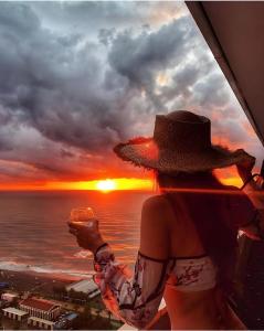PANORAMA Orbi Beach Resort Center Suite في باتومي: امرأة ترتدي قبعة تحمل كأساً من النبيذ وتطل على غروب الشمس
