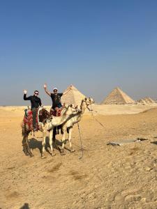 two men riding on camels in the desert with pyramids in the background at Nana Pyramids Guest House in Cairo