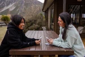 deux femmes assises sur une table de pique-nique en regardant leurs téléphones portables dans l'établissement Haka House Aoraki Mt Cook, à Mount Cook Village