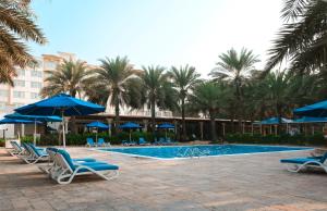 a pool with chairs and blue umbrellas and palm trees at Coral Beach Resort Sharjah in Sharjah