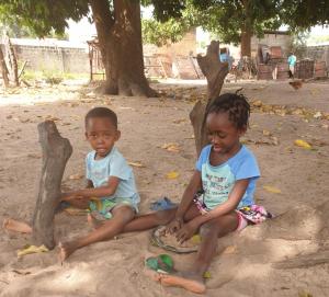 dwoje małych dzieci siedzących na piasku pod drzewem w obiekcie Live with the locals in Bamboo House w mieście Tanji