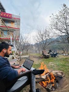 un hombre sentado en un banco junto a un fuego con un portátil en Burwa Breeze By Winsum Stays, en Palchān