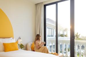 ホイアンにあるMoodhoian Riverside Resort & Spaの窓の外を見下ろすホテルの部屋の椅子に座っている女性