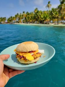 une main tenant une assiette avec un sandwich petit-déjeuner sur une plage dans l'établissement San Blas Sailing Experience With Us!, à El Porvenir