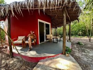 un homme assis sur une chaise devant une maison rouge dans l'établissement A Maze Inn Cottage, à Phumĭ Chroŭy Svay