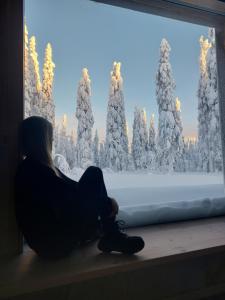a woman sitting on a window sill looking at a snow covered forest at Arctic Nest Forest Cabin in Kotila