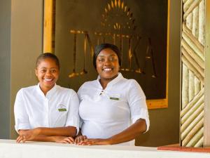 dos mujeres en camisas blancas de pie junto a una mesa en Insika lodge, en Victoria Falls