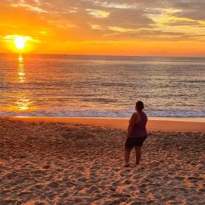 una mujer de pie en la playa mirando la puesta de sol en Suítes Som do Mar, en Caraguatatuba
