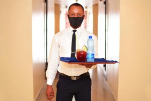 Ein Mann mit einer Maske, der ein Tablett mit einem Apfel und einer Flasche hält. in der Unterkunft Hotel Presidential in Port Harcourt