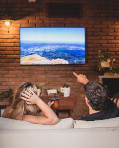 Ein Mann und eine Frau sitzen auf einem Stuhl und sehen einen Fernseher in der Unterkunft Hostel Falucho in Capilla del Monte