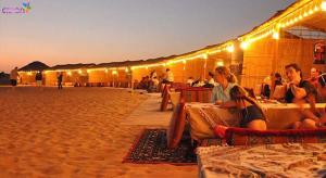 Enjoy The Leisure of Overnight Campsite in Dubai Desert Safari With Complementary Pick up في دبي: مجموعة من الناس يجلسون على الطاولات على الشاطئ