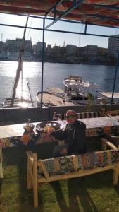 Un uomo seduto su una panchina di fronte a una barca di Onaty Narty Guest house a Aswan