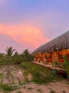 un bâtiment avec un toit de paille et un coucher de soleil en arrière-plan dans l'établissement Farmstay Sokfarm, à Trà Vinh