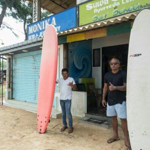 dos hombres parados frente a una tienda con una tabla de surf en Moragalla Beach Apartments, en Moragalla