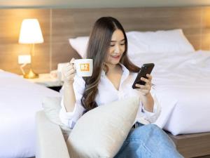 una mujer sentada en una cama sosteniendo una taza y un celular en B2 Lampang Boutique & Budget Hotel en Ban Nam Thong