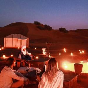Desert Luxury Camp Experience في مرزوقة: مجموعة من الناس يجلسون حول طاولة في الصحراء