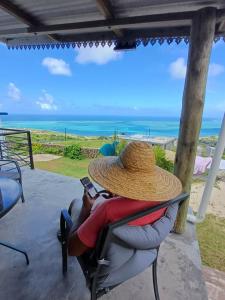 a person wearing a straw hat sitting in a chair looking at the ocean at Ô Bercail du Sud-Hibiscus chez Jeannette in Mt Bois Noir