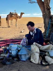 człowiek siedzący pod drzewem z wielbłądem w tle w obiekcie Adventure dans le desert w mieście Tagounite