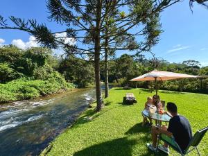 a family sitting at a table under an umbrella next to a river at Pousada Chalés Jardim das Bromélias - Visconde de Mauá in Visconde De Maua