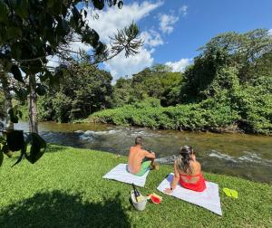 a man and a woman sitting on the grass by a river at Pousada Chalés Jardim das Bromélias - Visconde de Mauá in Visconde De Maua