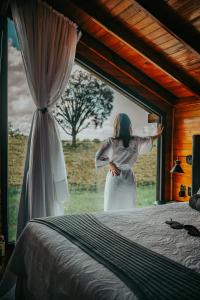 a woman standing in front of a window next to a bed at Pousada Capanna del Vale - Vale dos Vinhedos in Bento Gonçalves