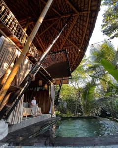 a woman sitting in a chair next to a house with a swimming pool at Eco Bamboo Island Bali - Bamboo House #4 in Selat