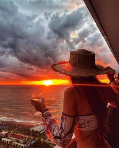 a woman in a hat holding a glass of wine looking at the sunset at PANORAMA ORBI City Center Suite Sea View in Batumi