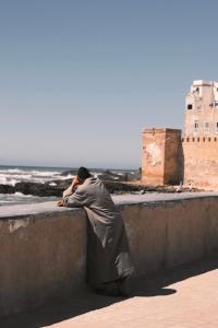 een man die op een muur zit en naar de oceaan kijkt bij Riad Bianca Essaouira Guest House in Essaouira