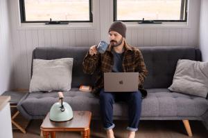 a man sitting on a couch holding a cell phone and a laptop at Odyssean Tiny House B in Cessnock