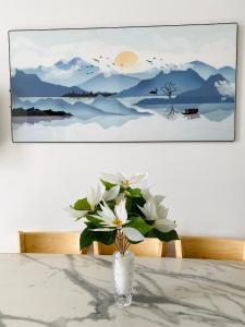 un vaso con fiori bianchi su un tavolo con un dipinto di ALPHA HOMESTAY MARINA 2 Phòng Ngủ View Sông a Ấp Ðông An (1)