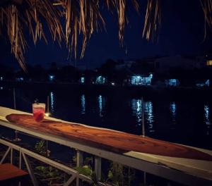 a drink sitting on a table next to the water at night at Here Library - A wooden home by the river with a library in Thốt Nốt