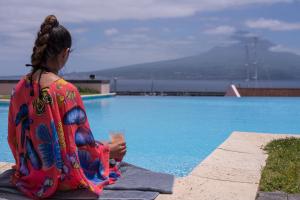 a woman sitting next to a swimming pool with a drink at Azoris Faial Garden – Resort Hotel in Horta