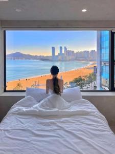 a woman sitting on a bed looking out a window at a beach at Paradise on earth in Busan