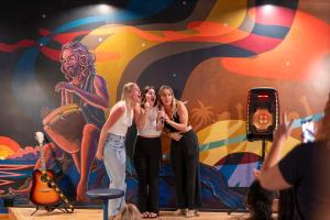 three women standing on a stage in front of a mural at Howzit Hostels in Wailuku
