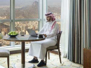 einem Mann, der an einem Tisch sitzt, mit einem Laptop vor einem Fenster in der Unterkunft Movenpick Makkah Hajar Tower in Mekka