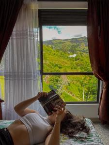 a woman laying on a bed reading a book in front of a window at Descanso Bajo La Piedra Del Peñol in Guatapé