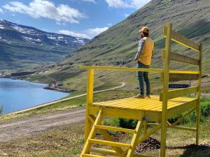 a man standing on top of a yellow chair overlooking a mountain at Langahlid Cottages & Hot Tubs in Seyðisfjörður
