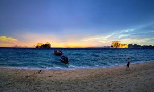 a man standing on a beach with a boat in the water at Thanya Beach Resort - SHA Plus in Ko Ngai