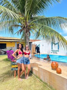 two women standing under a palm tree next to a swimming pool at Dr House veraneio Abaís praia SERGIPE in Praia Da Caueira +6 photos