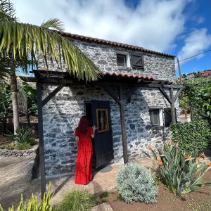 Eine Frau in einem roten Kleid steht vor einem Steinhaus in der Unterkunft As Casinhas da Madeira - Cosy Stone Cottage in a subtropical garden in Arco da Calheta