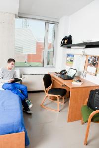 un homme assis sur un lit dans une pièce avec un bureau dans l'établissement University of Toronto-New College Residence-45 Willcocks Residence, à Toronto