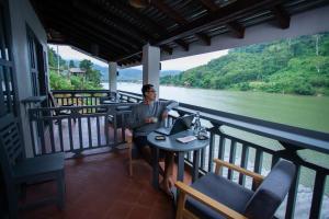 a man sitting at a table with a laptop on a balcony at Dokchampa River View Villa in Nongkhiaw