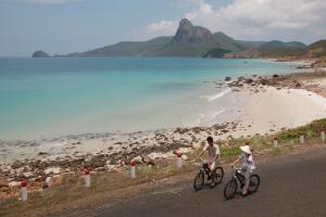 two people riding bikes on a road next to a beach at Six Senses Con Dao - Turtle Island Paradise in Con Dao