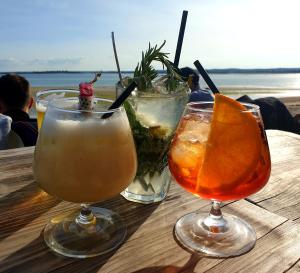 three drinks on a wooden table with a view of the ocean at Gîte du Clos Giot in Saint-Vaast-la-Hougue