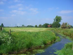 a river in a field with grass and windmills at Landhaus am Kleeweg in Osteel