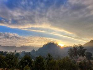 a misty sunrise in the mountains with palm trees at มาค่ะ ฟาร์มสเตย์ ห้วยเขย่ง สวนคุณตา สวนทุเรียนทองผาภูมิ in Ban Diso