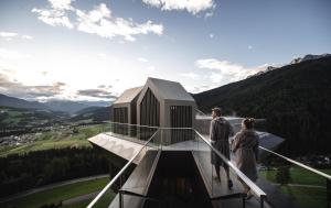 due persone in piedi sopra una torre di vetro con un edificio di Alpin Panorama Hotel Hubertus a Valdaora
